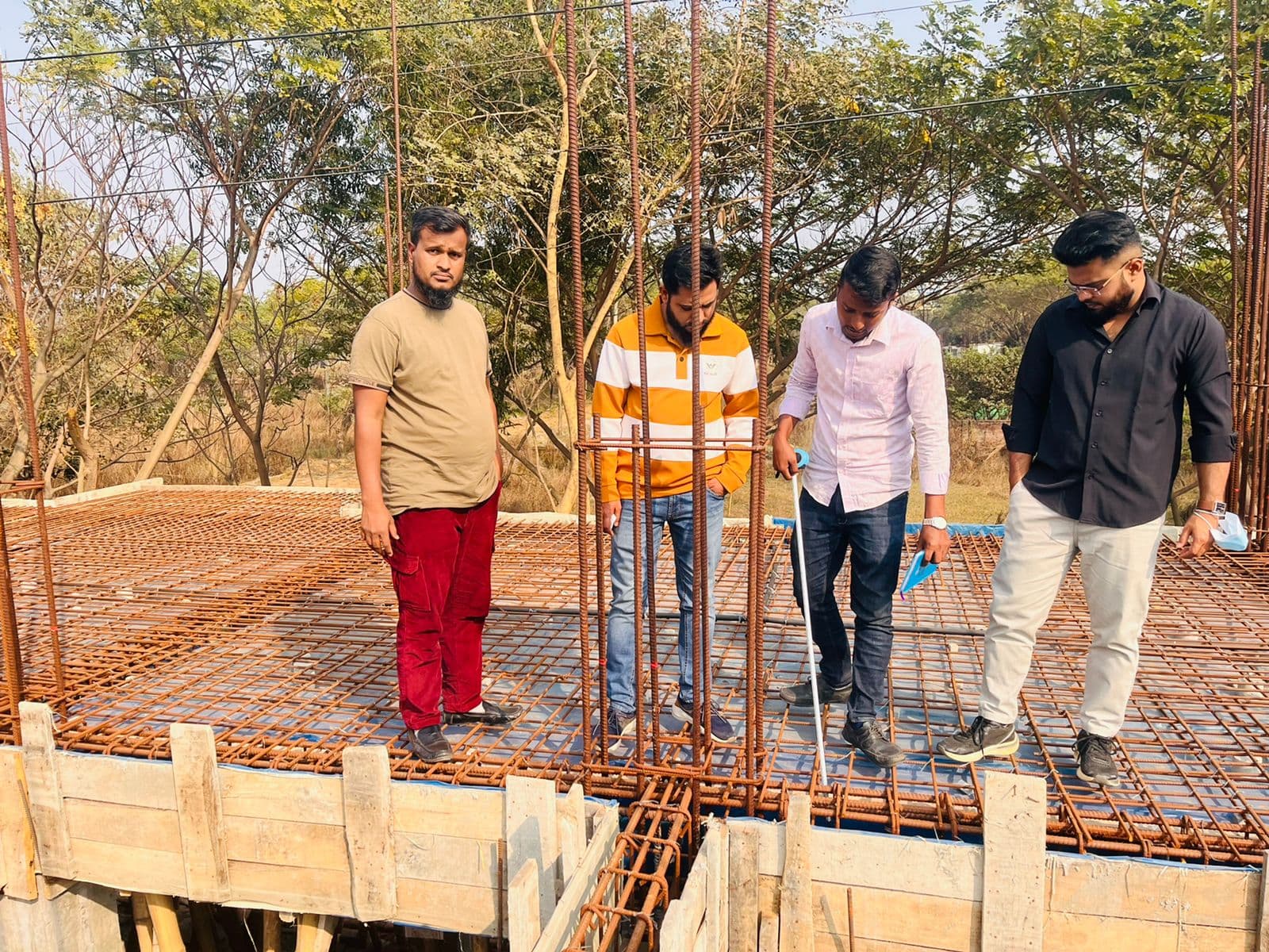 Four men in casual and business dress standing on a rebar-reinforced concrete floor slab awaiting a pour, timber shuttering at the edge, daylight