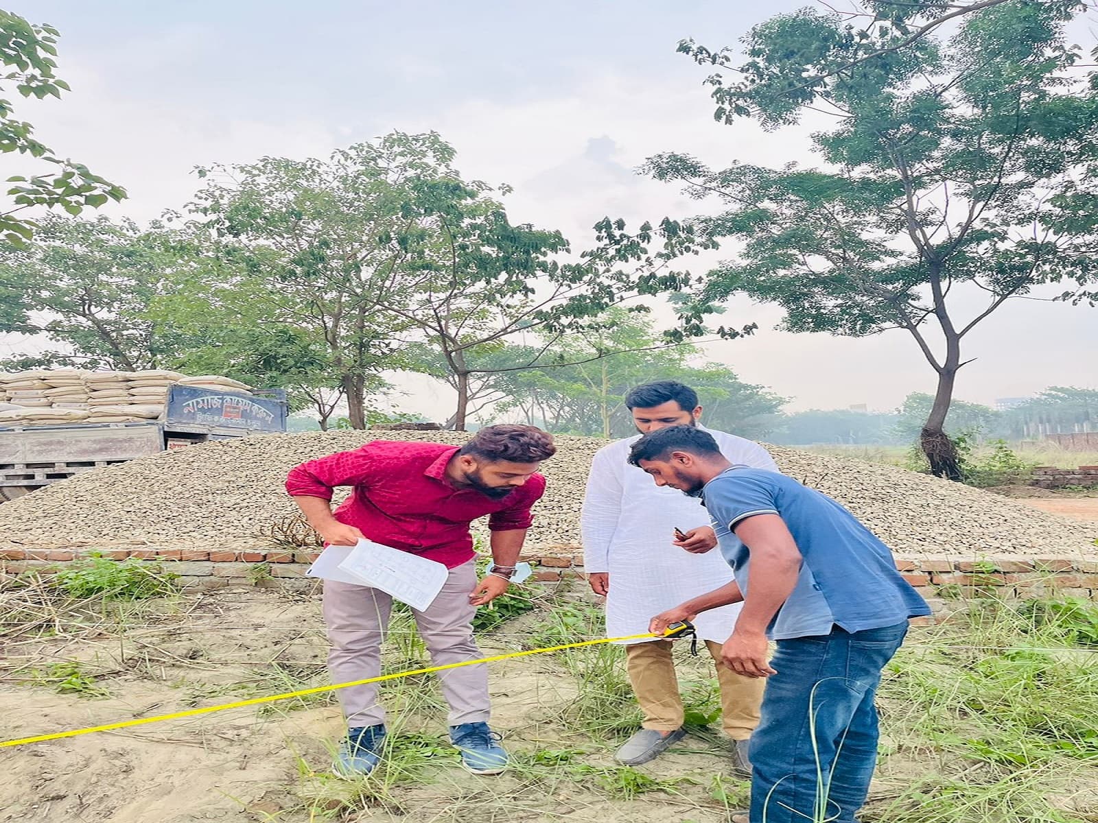 Three men in an open field beside piles of stone aggregate and a loaded truck, one holding site drawings while another stretches a measuring tape
