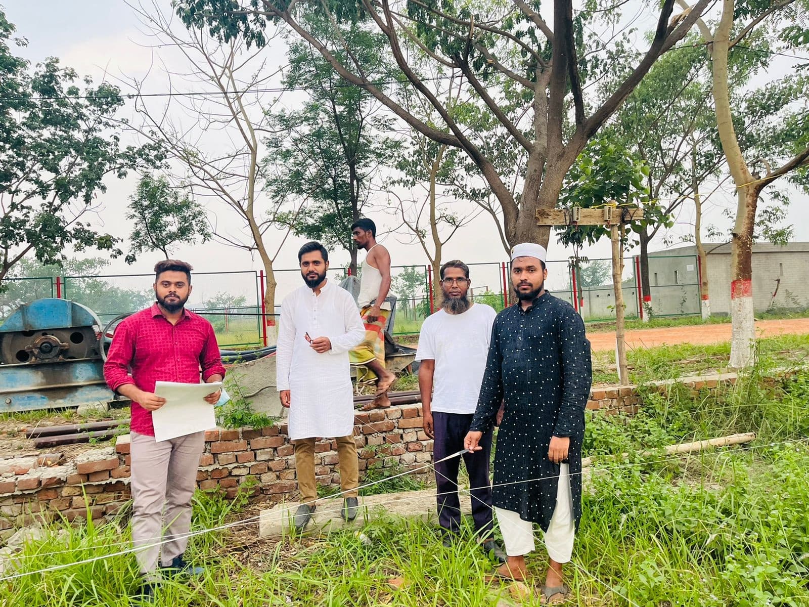 Team members standing at a rural plot boundary marked out with string lines and low brick stub walls, trees and a labourer in the background