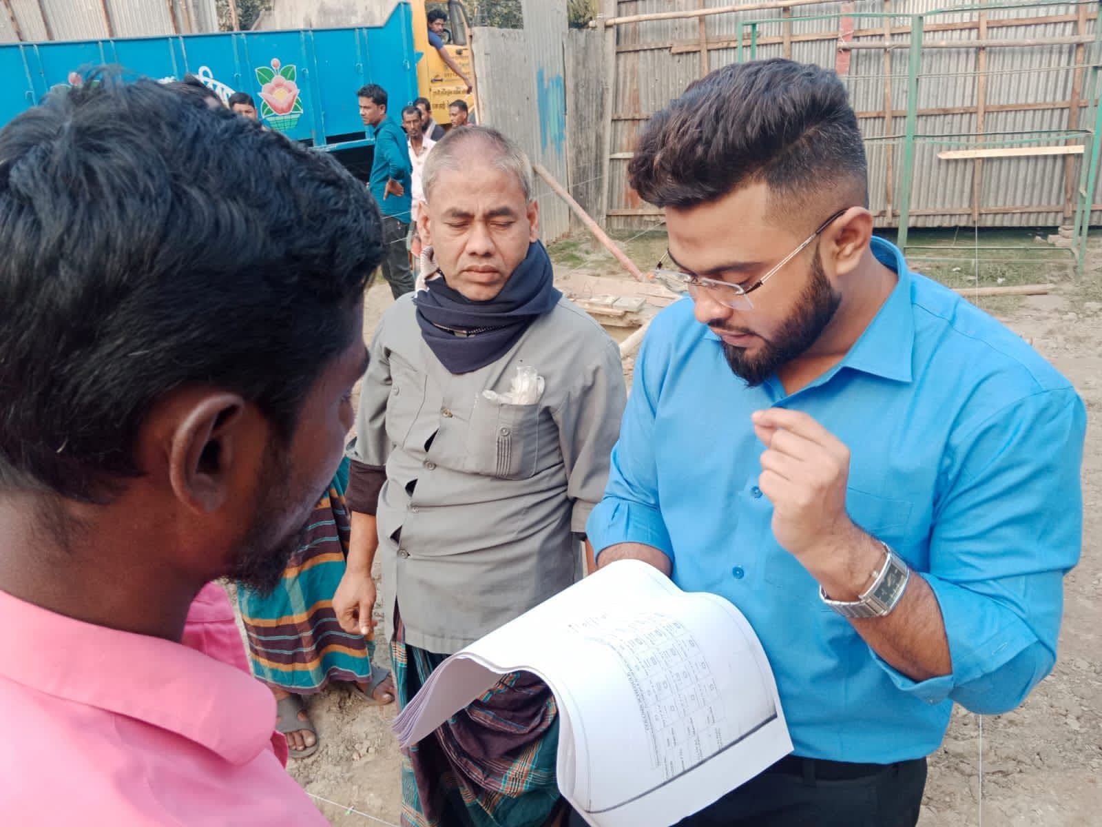 Close view of a Kashban staff member reviewing a bound site document with two other men at a corrugated-tin site boundary, onlookers behind