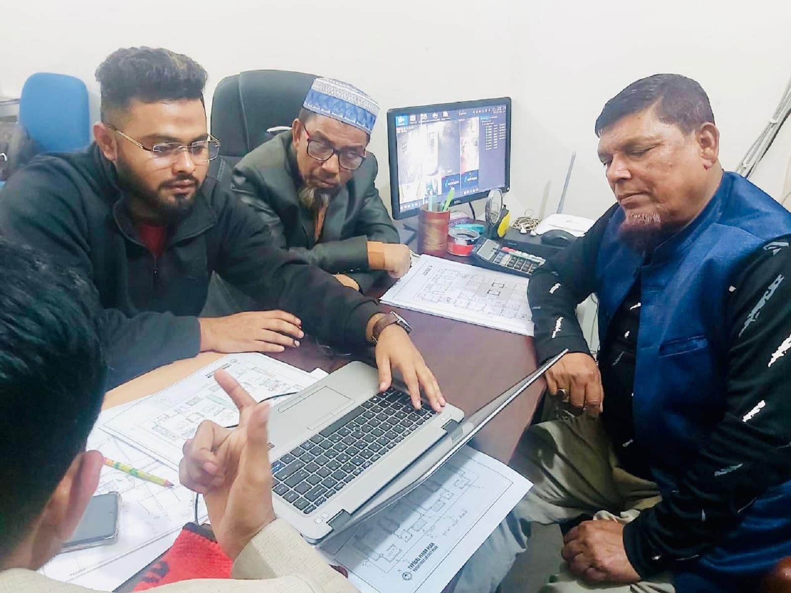 Four men gathered around a laptop and printed floor plans at an office desk, a monitor behind them showing split-screen site camera feeds
