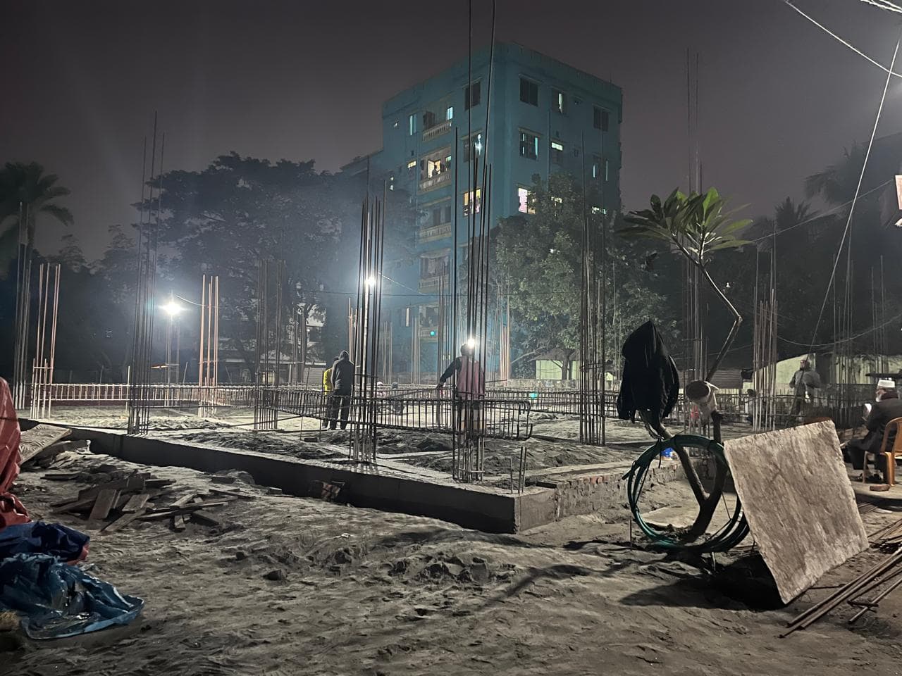 Night pour in Mohammadpur: bare reinforcement cages standing in a sand-filled foundation plot under two work lights, a pale blue-lit apartment block behind