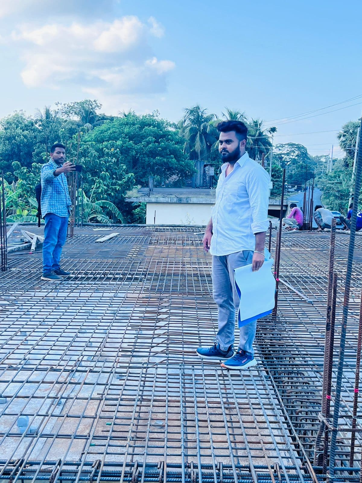 Daytime rooftop-slab photo: dense steel rebar mesh underfoot, one man in white holding site papers while another photographs the view on his phone
