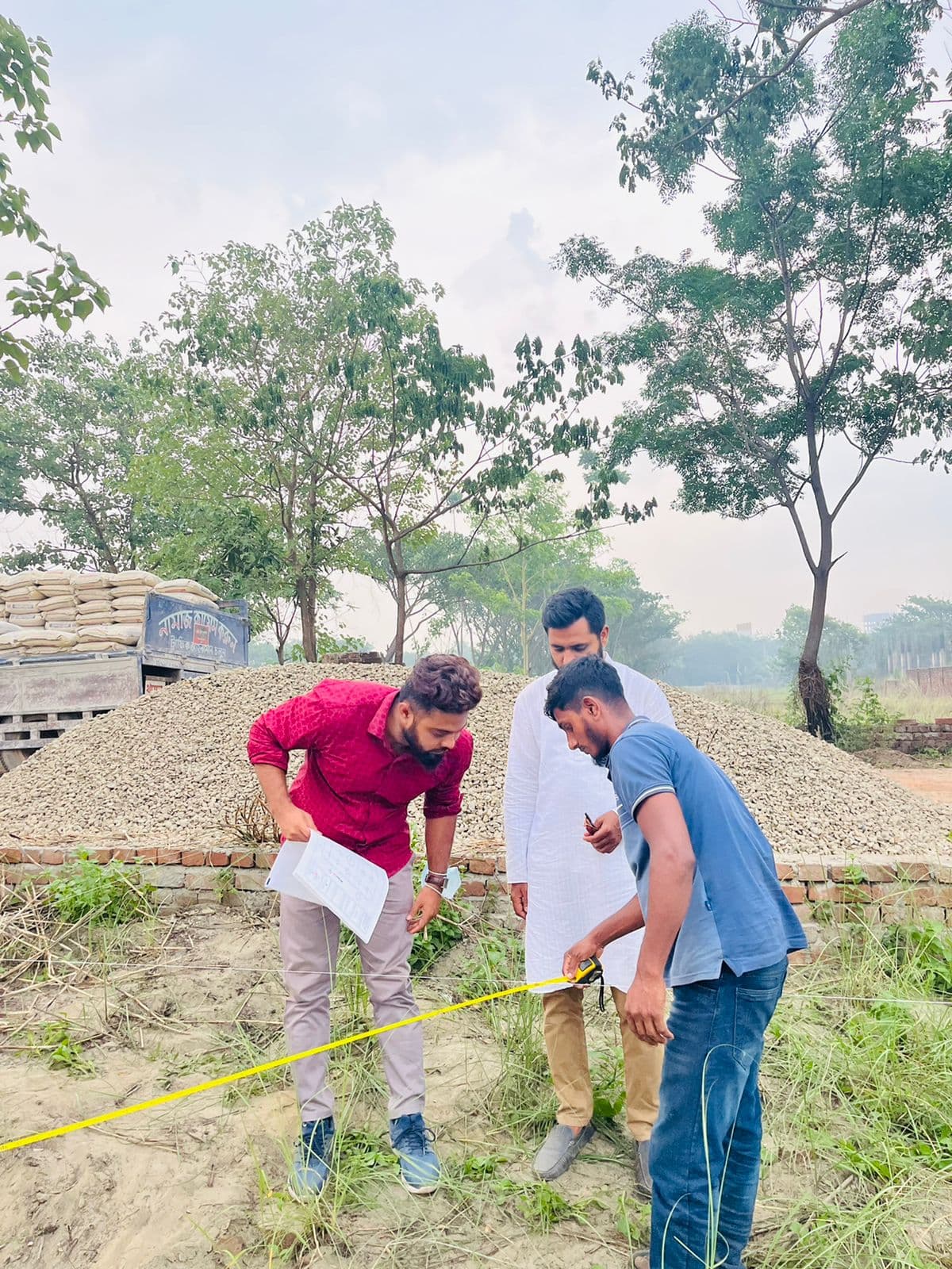 Field survey scene: men measuring a plot with a tape line beside piles of stone aggregate and a loaded delivery truck, overcast sky