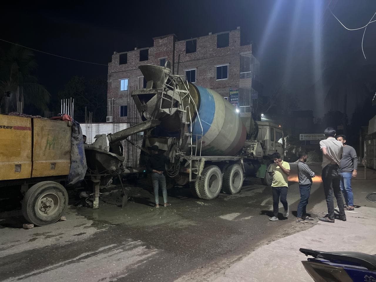 Night concrete pour: a mixer truck feeding a pump beside a partially built brick building with lit upper-storey windows, workers watching from the street